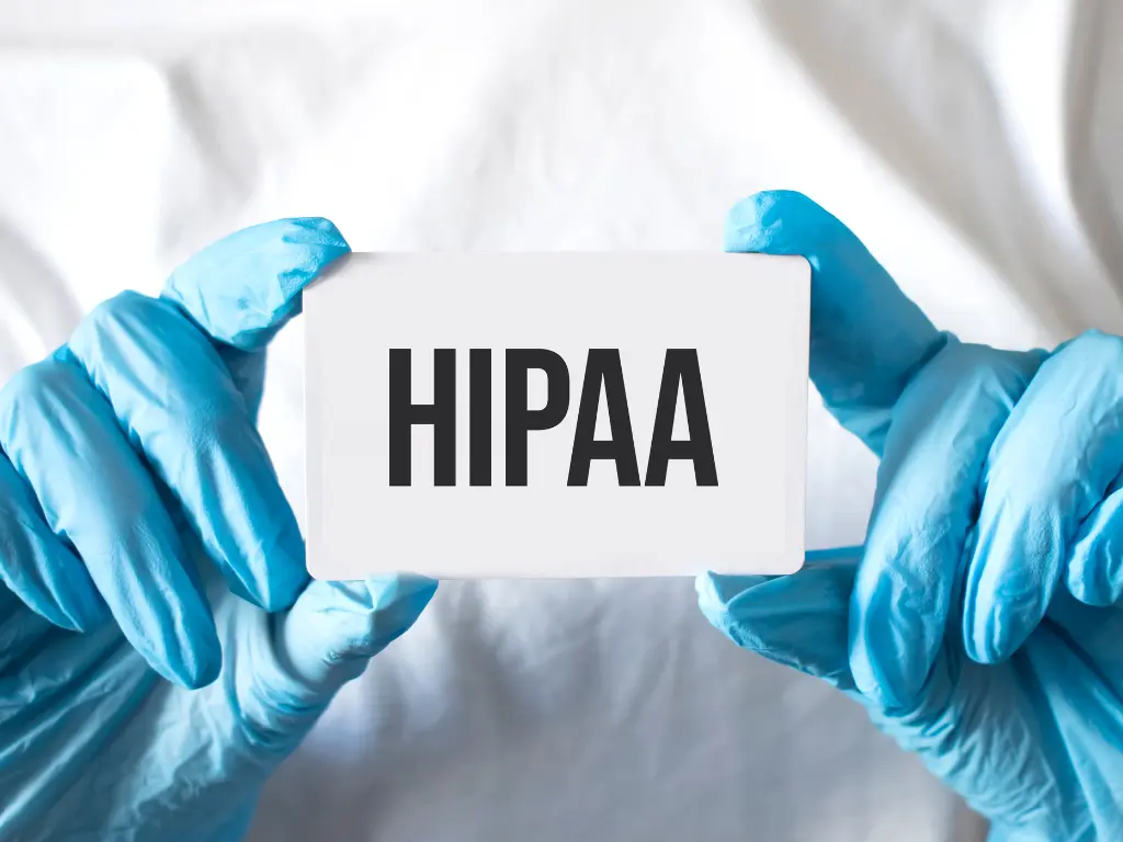 Person wearing blue medical gloves holds a card with the word "HIPAA" printed in bold black letters.