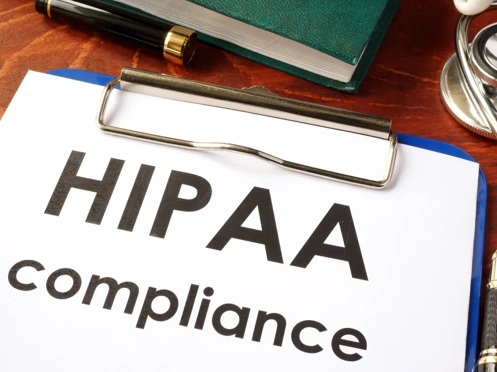 A clipboard with a document labeled "HIPAA Compliance" on a desk, along with a green folder and pen.