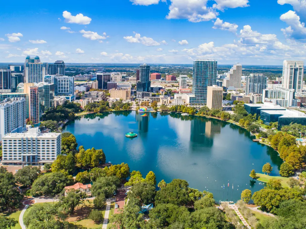 Aerial view of a city skyline with a large, round lake surrounded by trees and tall buildings under a blue sky with scattered clouds.