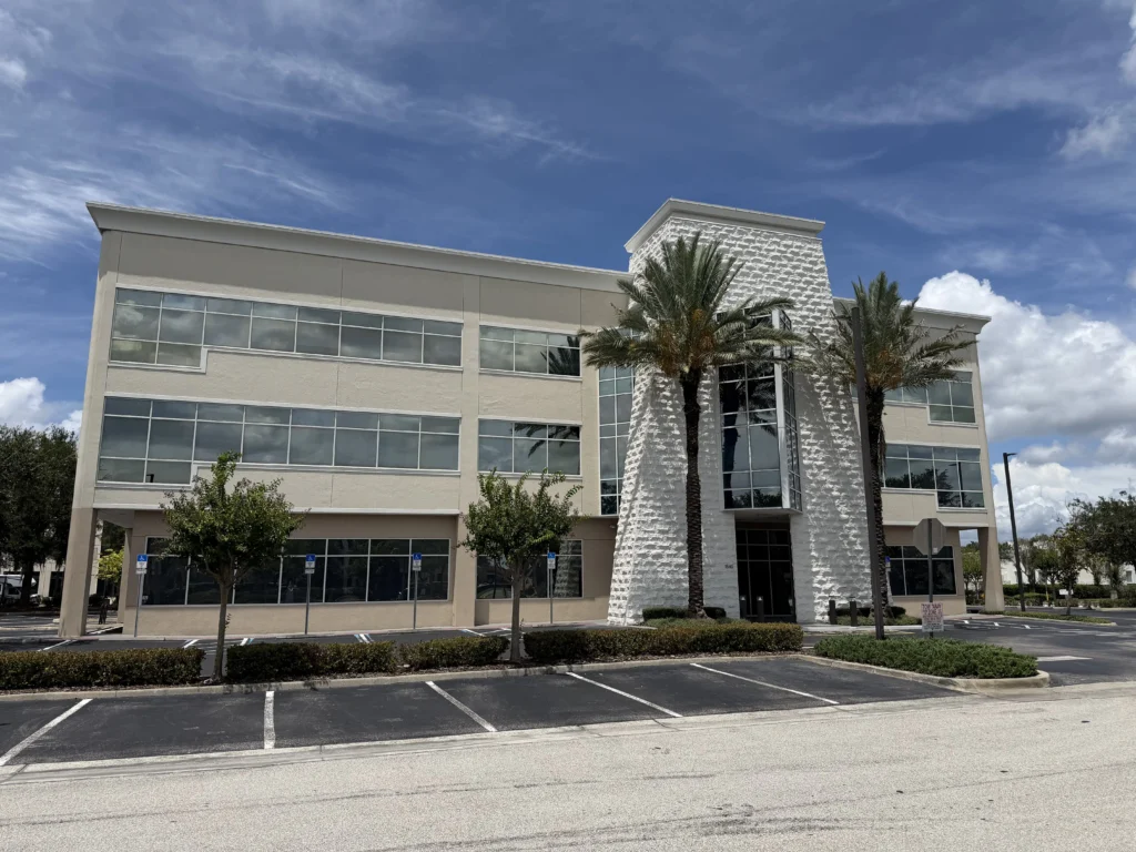 Modern three-story office building with large windows, palm trees, and a parking lot in front — the Harmony MSP offices in Lake Mary, FL.