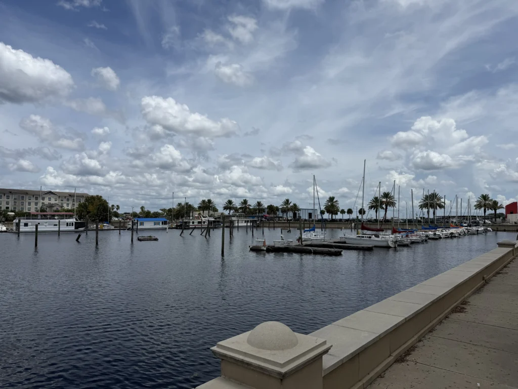 A marina with docked sailboats, calm water, palm trees, and buildings under a partly cloudy sky.