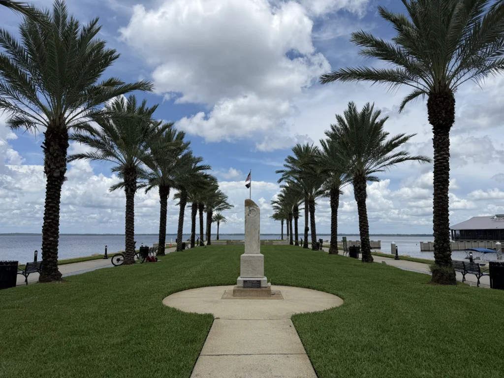 A walkway lined with palm trees, leading to a monument and flag, next to a waterfront under a cloudy sky.