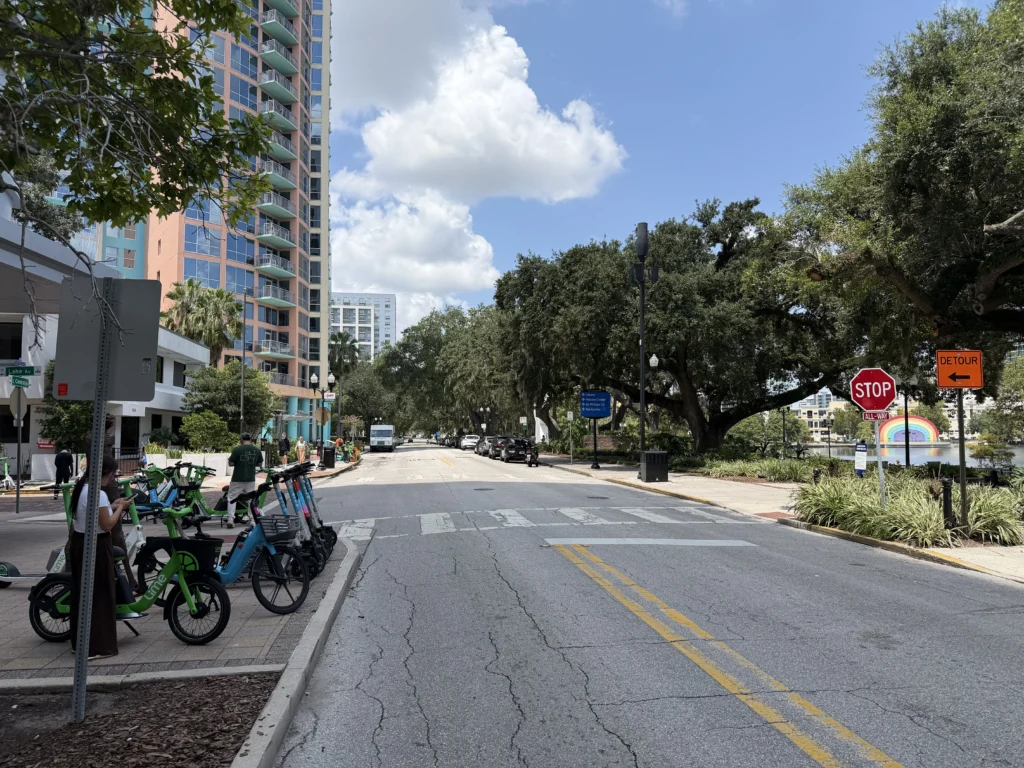 Urban street with bike and scooter rentals, tall buildings on the left, trees lining the right, stop and detour signs, and a rainbow sculpture near a lake in the background.