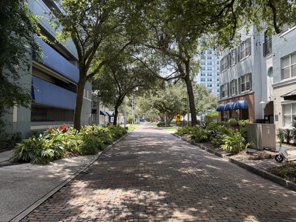 Tree-lined brick street flanked by modern buildings, lush green plants, and deep shade from large overhead trees on a sunny day.