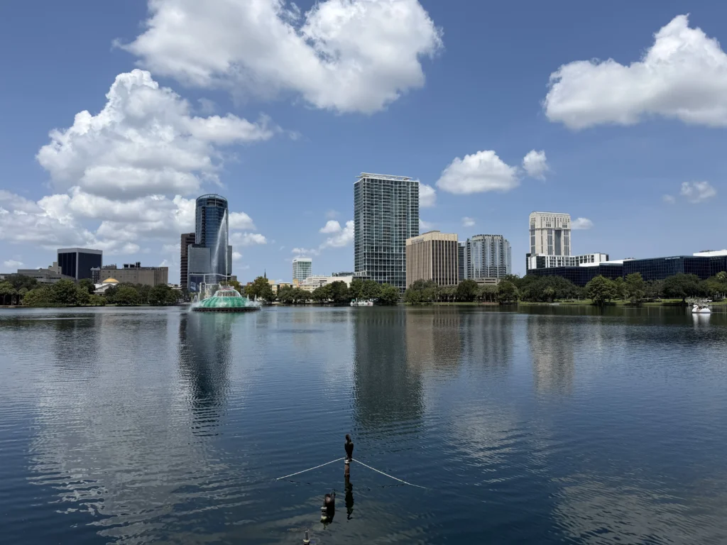 City skyline with tall buildings reflecting on a lake, a green fountain in the water, partly cloudy blue sky above.