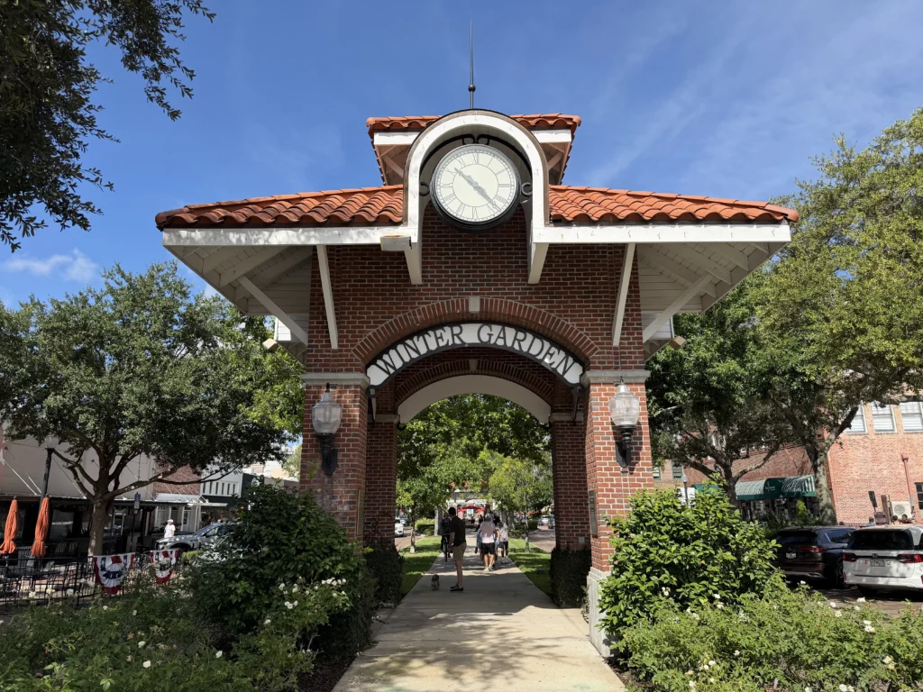 A red-brick archway with a clock on top, surrounded by green bushes and trees, leads to a park on a sunny day.