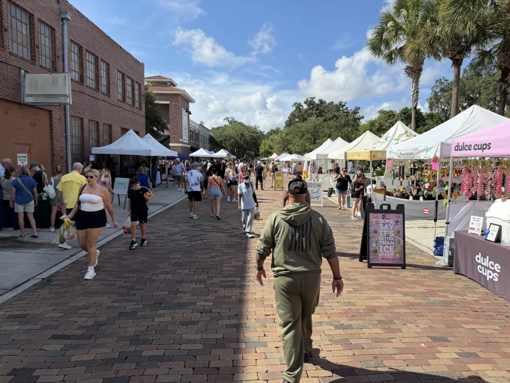 People walking along a brick street lined with white vendor tents at an outdoor market on a sunny day.