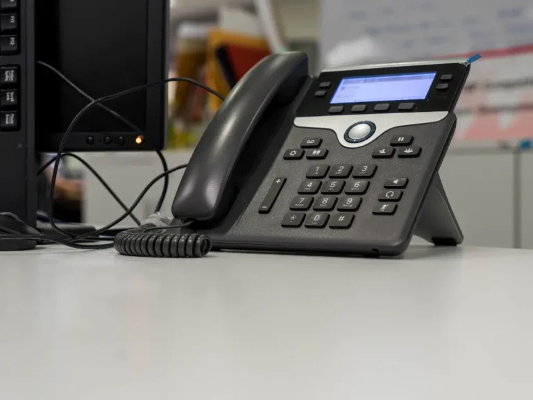 A black office desk phone with a digital display and buttons sits on a white desk next to a computer monitor.