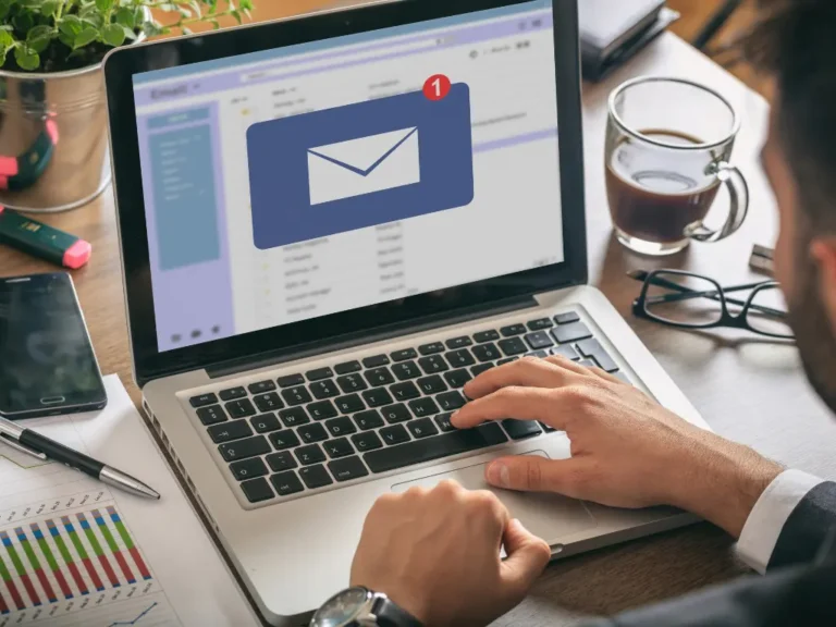Person checking email on laptop with a new message notification, sitting at a desk with a coffee cup, graphs, pen, smartphone, and glasses.