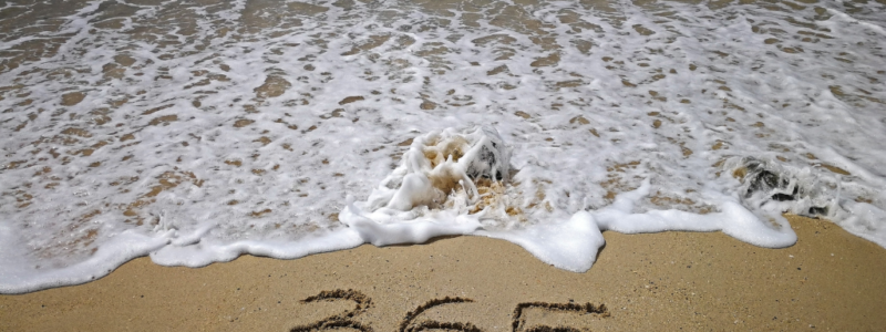 The number 365 is written in the sand on a beach, with ocean waves approaching the shoreline.