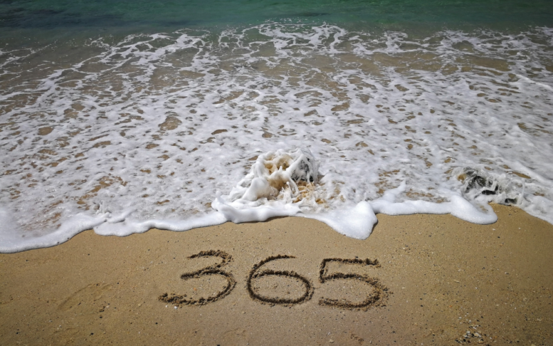The number 365 is written in the sand on a beach, with ocean waves approaching the shoreline.