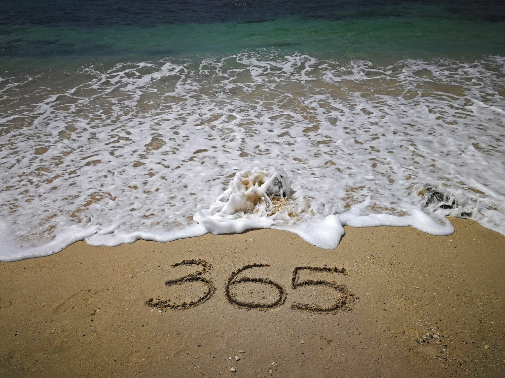 The number 365 is written in the sand on a beach, with ocean waves approaching the shoreline.