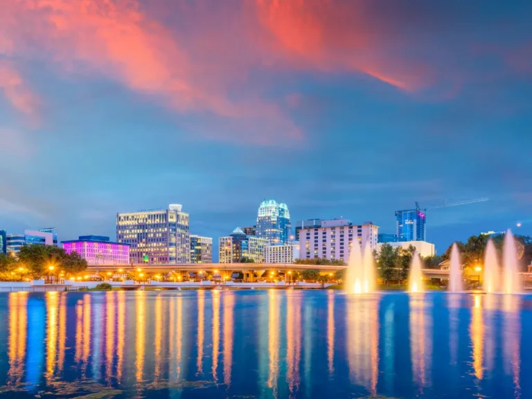 Orlando skyline at sunset with colorful buildings, glowing lights, water fountains, and vibrant reflections on a calm lake under a dramatic pink and blue sky.