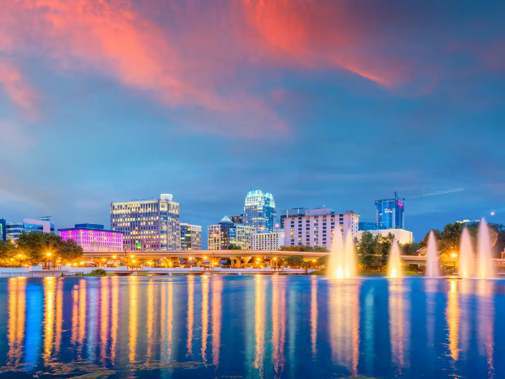 Orlando skyline at sunset with colorful buildings, glowing lights, water fountains, and vibrant reflections on a calm lake under a dramatic pink and blue sky.