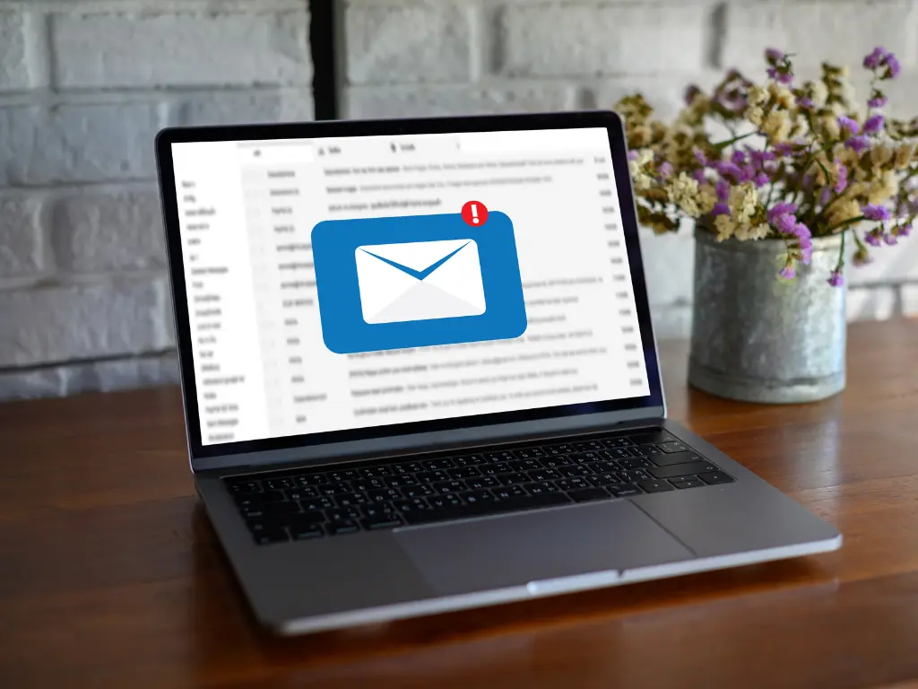 A laptop displaying an email inbox with a new message notification icon, placed on a wooden table next to a bucket of flowers.