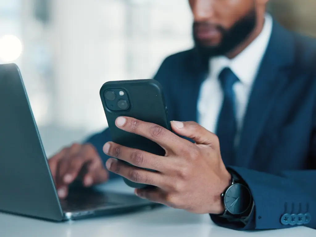 A man in a suit using a laptop and holding a smartphone, focusing on multitasking in a professional workspace.