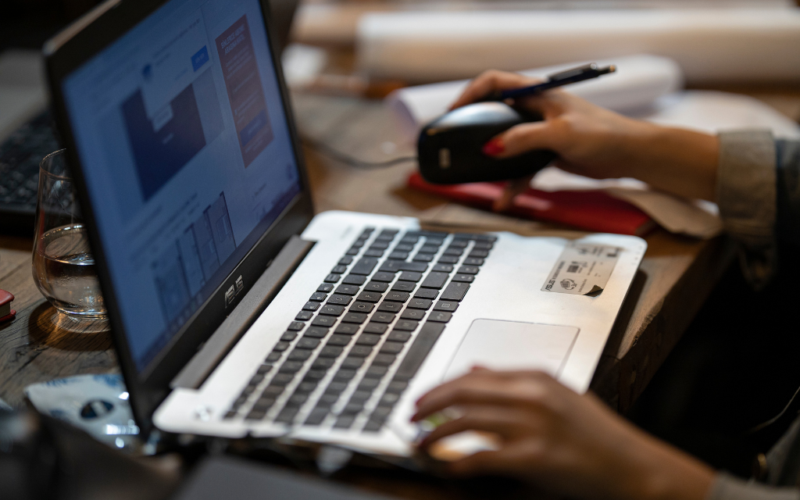 Close-up of a person using a laptop and mouse at a desk, with papers, a pen, and a glass of water nearby.