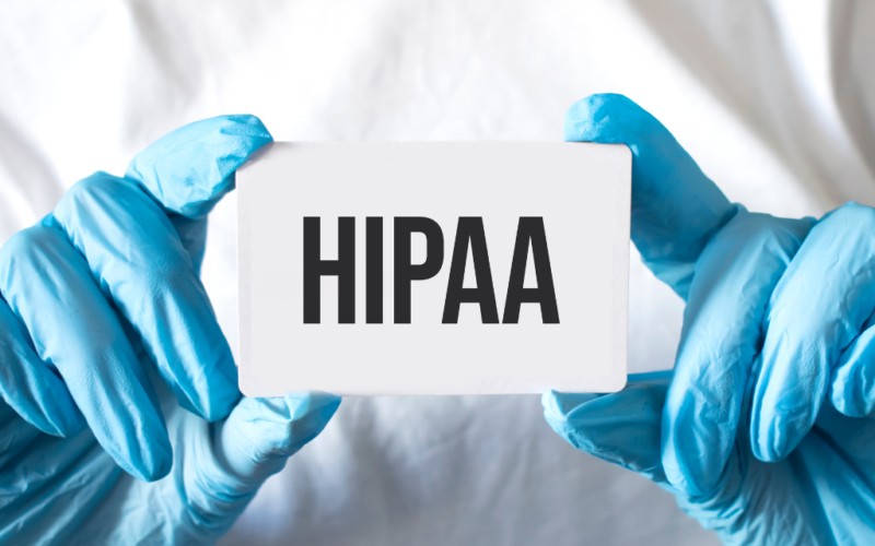 Person wearing blue medical gloves holds a white card with the word "HIPAA" printed in bold black letters.