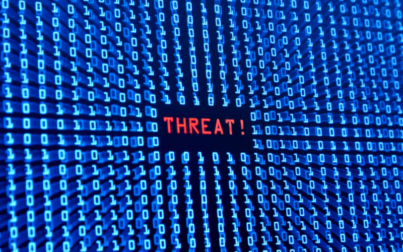 Rows of blue binary code surround the red word "THREAT!" on a dark digital screen, symbolizing a cybersecurity or computer virus warning.