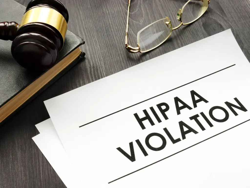 A gavel, eyeglasses, and a document labeled "HIPAA Violation" on a dark wood desk, symbolizing legal or privacy issues.