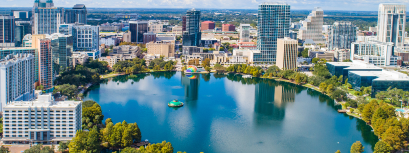 Aerial view of the Orlando skyline with tall buildings surrounding Lake Eola and green park under a blue sky with clouds.