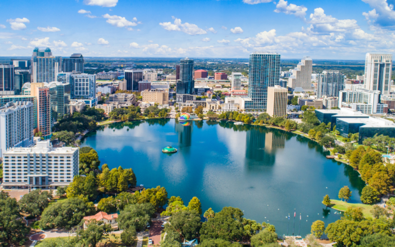 Aerial view of the Orlando skyline with tall buildings surrounding Lake Eola and green park under a blue sky with clouds.