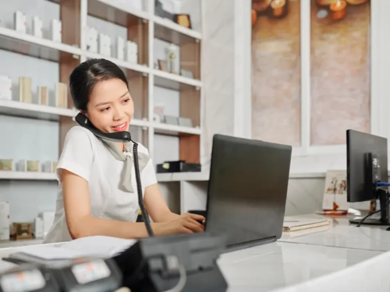 A woman at a desk multitasks by talking on the phone and typing on a laptop in a modern office setting.