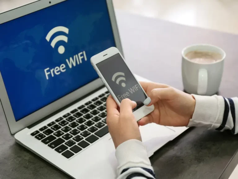 A person holding a smartphone, both laptop and phone screens display "Free WIFI," with a cup of coffee on the table beside them.