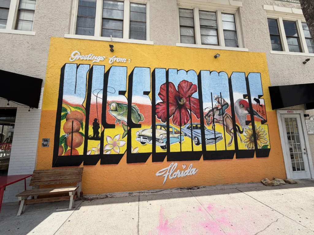 Colorful mural reads “Greetings from Kissimmee Florida,” featuring local icons like oranges, flowers, a fish, birds, a cowboy, and classic cars on a yellow-orange wall.