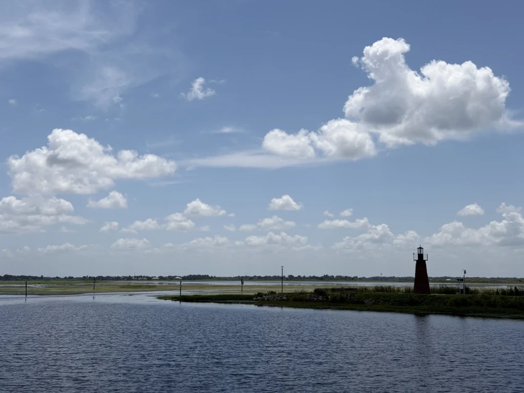 A small lighthouse stands by the water under a bright blue sky with scattered white clouds.