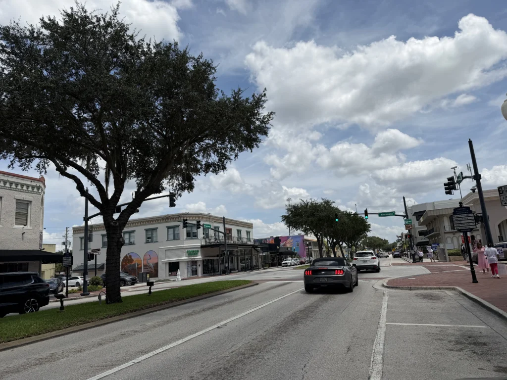 A city street with cars driving, pedestrians on sidewalks, trees lining the road, historic buildings, and a partly cloudy sky overhead.