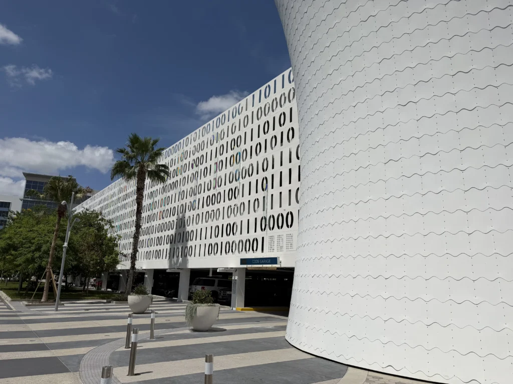 Modern building with circular patterned facade, palm trees, and a striped sidewalk under a clear blue sky.
