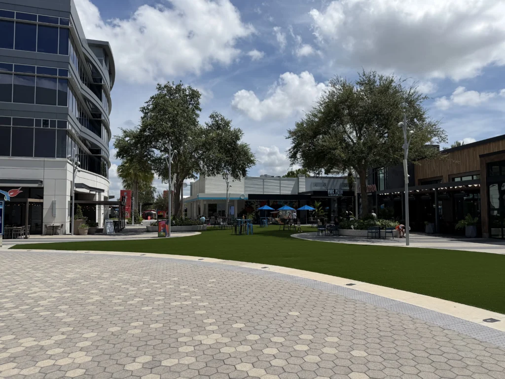 Modern outdoor plaza with hexagonal paving, green grass area, trees, and surrounding restaurants and shops under a partly cloudy blue sky.