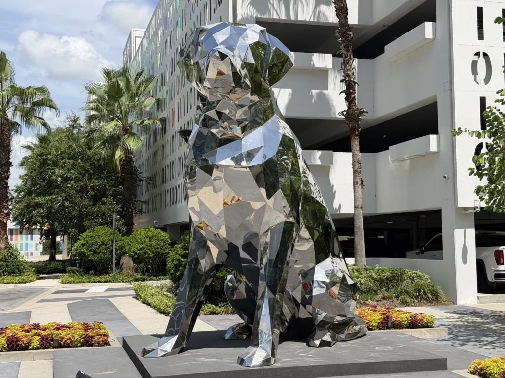 Large metallic geometric dog sculpture sits outdoors near palm trees and a multi-story parking garage, reflecting light and nearby plants.