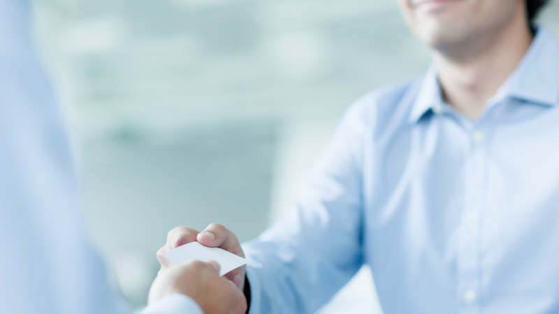 Two people in business attire exchanging a business card in a bright, professional setting.