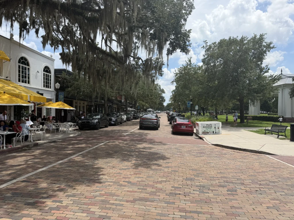 A brick street lined with parked cars, outdoor café seating with yellow umbrellas, and large oak trees with Spanish moss under a partly cloudy sky.